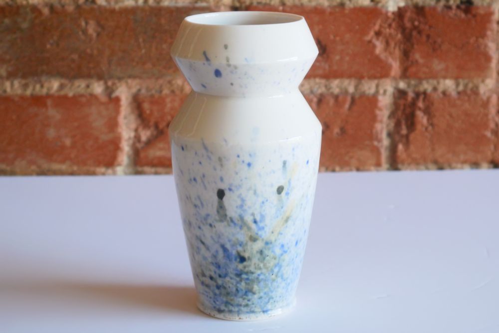 A shiny white vase with splatters of blues and greens that seem to drip from the base upward. It has a narrow foot, and slightly rounded walls out to a wider shoulder – then cuts in sharply, out again to the same width, and back in – almost like a collapsible accordion at the top