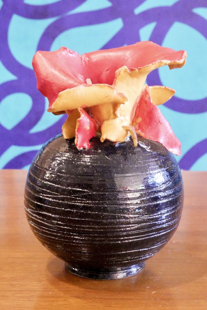 The bottom of the vase is a shiny black orb on a shallow pedestal, with uneven grooves carved around the surface. up top, slabs of torn clay explode out in satin-semi-gloss red and yellow leaves or pedals that almost form a flower, or almost the flared neck of a vase. There is nothing that makes the two parts cohere in a single piece. 