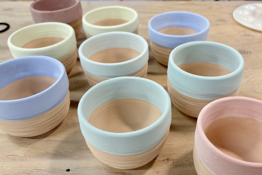 nine uniform small round-bottom planters with dry unfired glazes around each rim in pastel shades of red and blue and green and purple