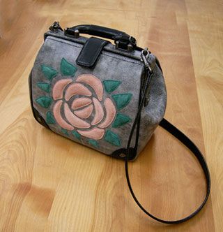 「さびしがりやのクニット」旅のカバン for the shop PIEKKO. Sitting on a hardwood floor, a grey stonewashed cowhide leather bag with a large pink and green large suede rose appliqué with raised texture. An adjustable shoulder-strap lays on the floor next to the bag. Produced for the shop PIEKKO at Dream Pocket.