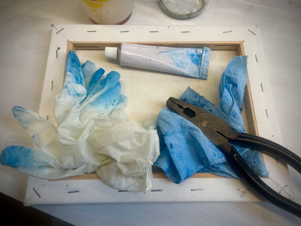 a photo of a paint tube, white vinyl gloves, paper towel, and pliers. Everything is covered in blue smudges