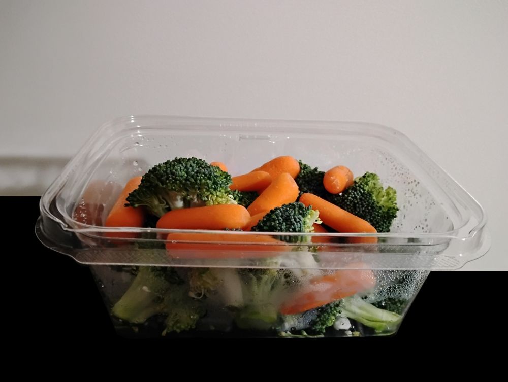 a picture shows a square plastic tray of vegetables containing 10 small carrots amongst two fistfulls of broccoli.