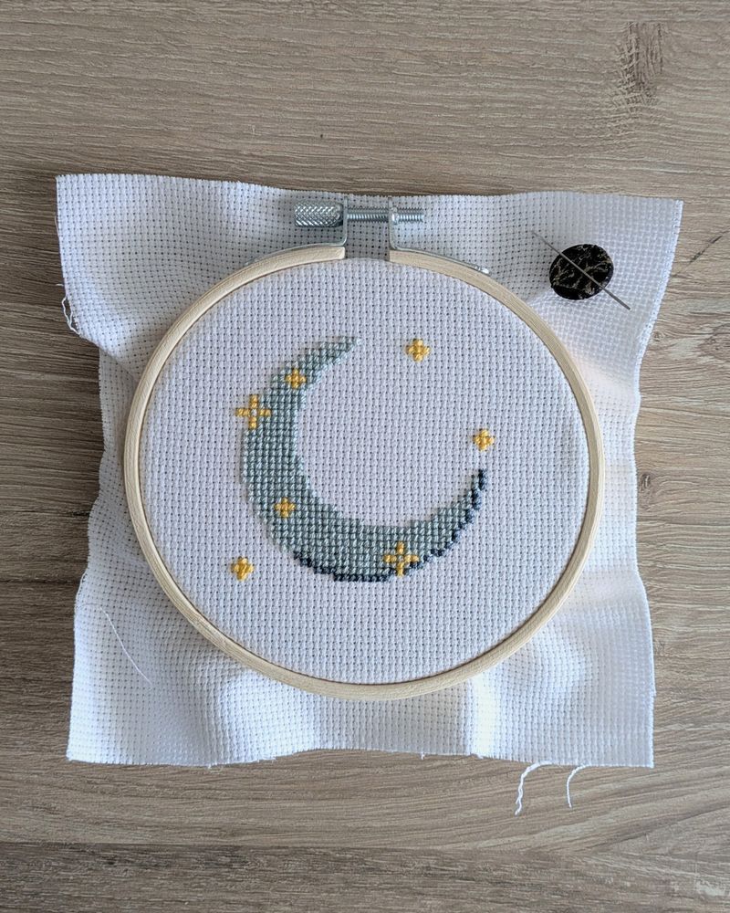 A WIP cross stitch in a 4-inch hoop with yellow sparkles and a crescent moon