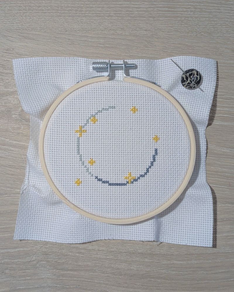 A WIP cross stitch in a 4-inch hoop with yellow sparkles and two shades of gray creating 3/4 of a circular line