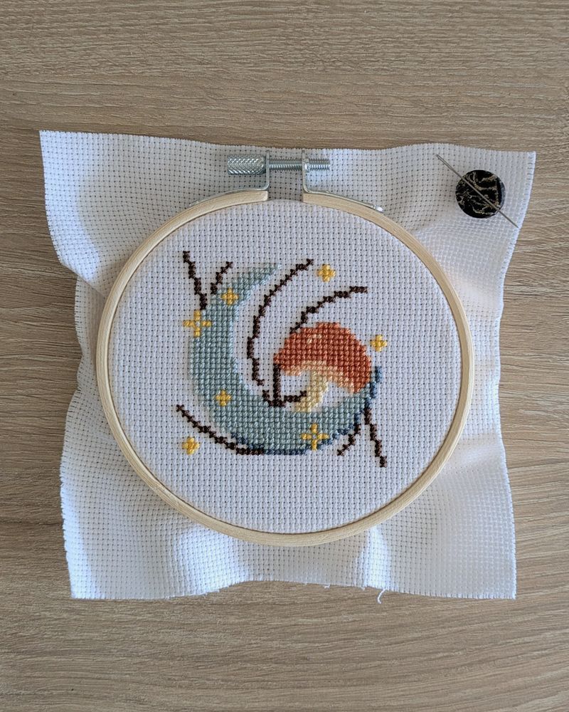 A WIP cross stitch of a gray crescent moon and an orange mushroom. Brown plant stems are stitched behind them.