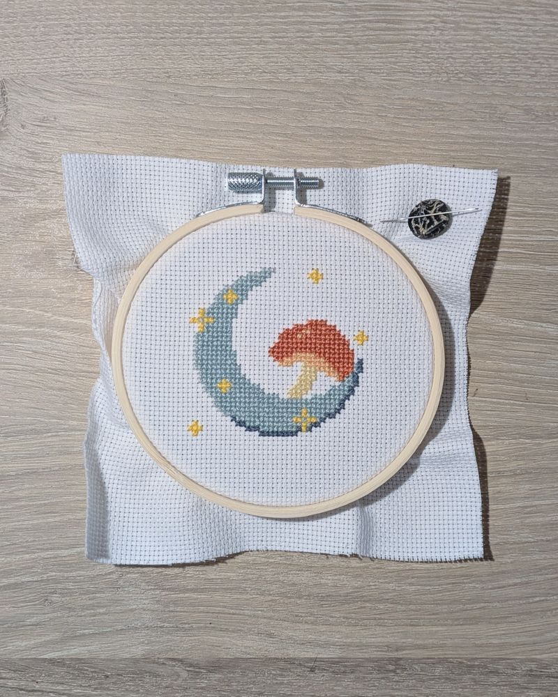 A WIP cross stitch in a 4-inch hoop with a gray crescent moon and an orange mushroom surrounded by yellow sparkles