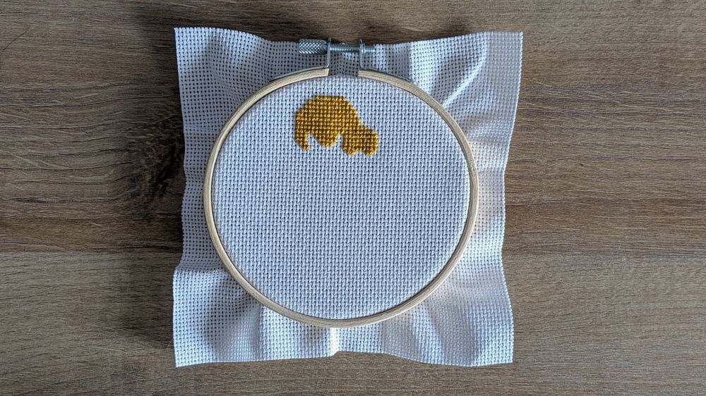An unfinished cross stitch on white fabric of the very top of Isabelle's head, the dog from Animal Crossing.