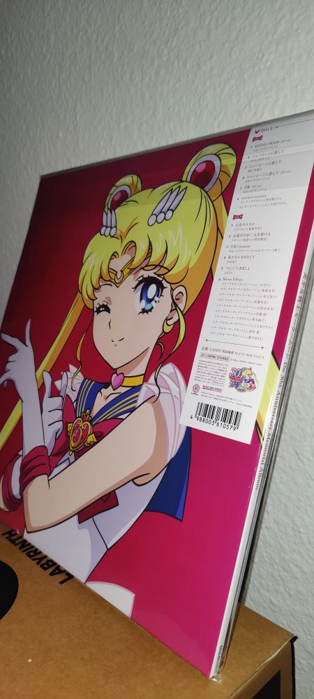 A close-up photo of a Sailor Moon 30th Anniversary vinyl record leaning against a wall. The cover features a bright, colorful illustration of Sailor Moon winking with her hand raised in a classic pose. A pink and red background enhances the vibrant design. A white tracklist label in Japanese is visible on the top-right corner of the packaging.
