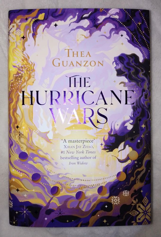 Cover of 'The Hurricane Wars' by Thea Guanzon. Is is in yellow and purple tones, the cover painted by Afterblossom. In the left side there is a woman in yellow and gold toned gown, on the right side is a man, cloaked in purple mists