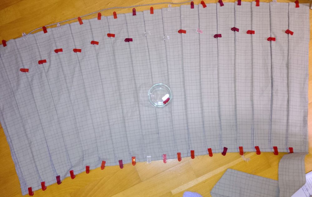 A piece of beige tartan patterned fabric lying on wooden floor boards. The fabric is pleated, the pleats held in place with red plastic sewing clips