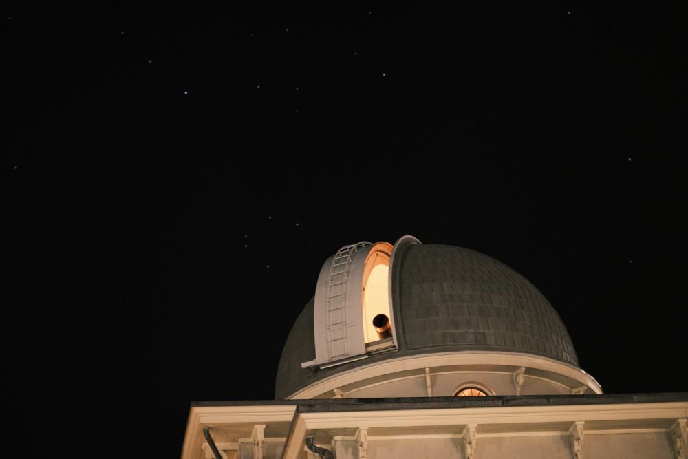 The Fitz refractor just barely visible through the dome slit of the small observatory dome made 170 years ago. Incandescent yellow orange light illuminating the inside of the building. The western night sky of fall is slightly detailed by a few stars. 