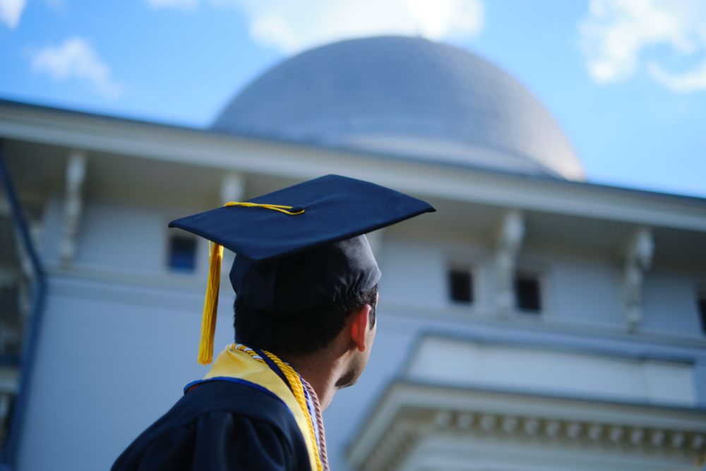 Person in graduation gown and cap looking up at dome of observatory. 