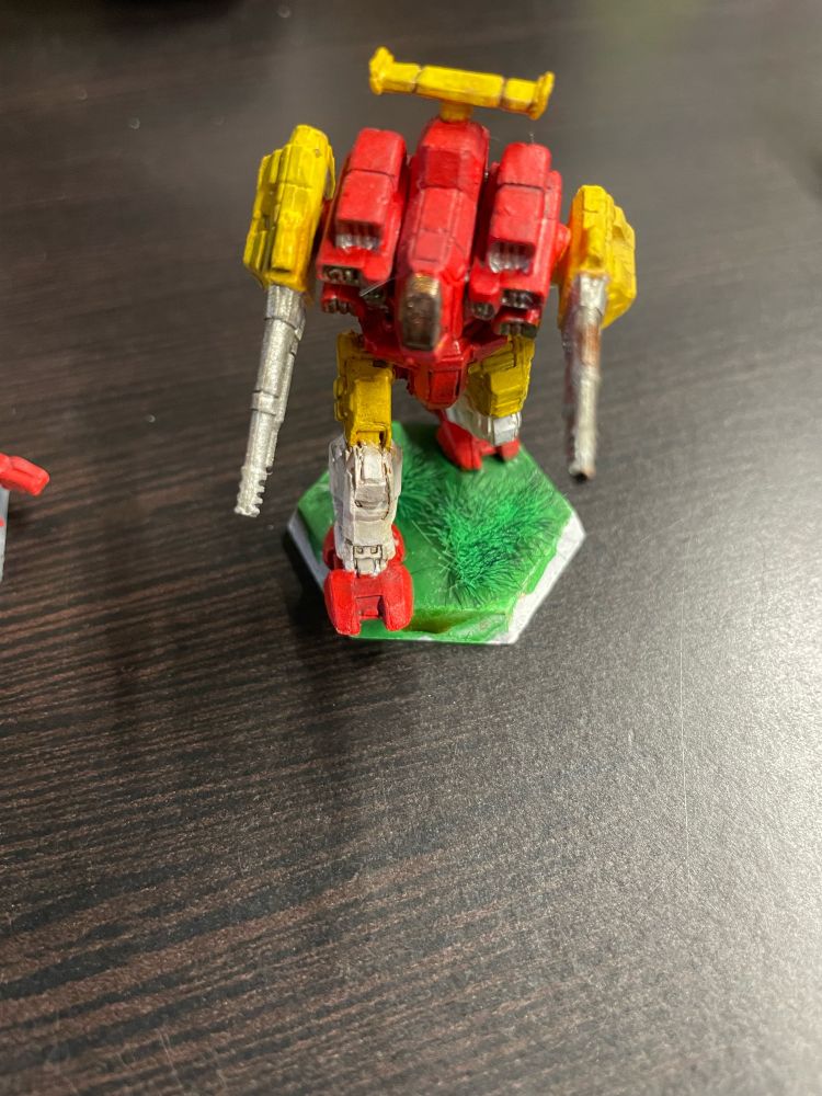 A Rifleman Battletech, with are torso and feet, yellow radar fin, upper arms, and upper legs, and white shins, with a cockpit in shiny red-gold like the F-22, and silvery highlights. The base is green with green grass tufts. He is like unto a Kansas City Chiefs player. 