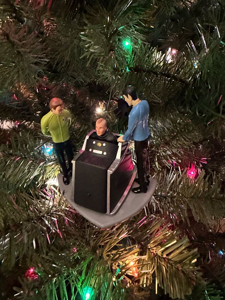 A Christmas tree with an ornament depicting Kirk and Spock standing with Captain Christopher Pike in “The Menagerie”, where he is in a futuristic wheelchair and can only communicate with beeps from the chair. What in the hell, man