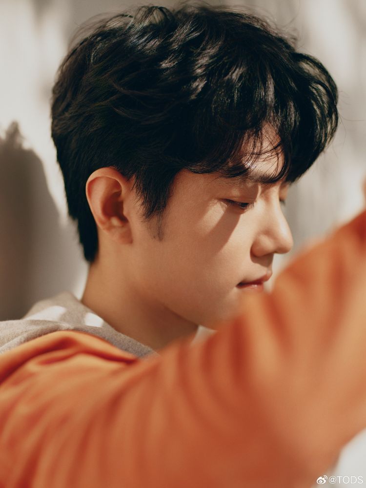Closeup of Xiao Zhan leaning to a wall with his eyes closed. He's wearing a orange jacket.