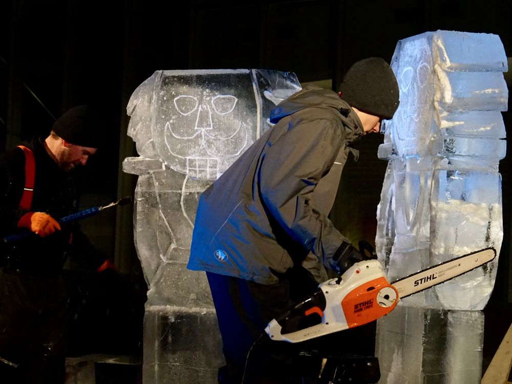 Two people carving ice sculptures. One of the people is using a chainsaw. Both are dressed for cool weather, but not cold. The sculptures are a little undefined but appear to be nutcrackers 
