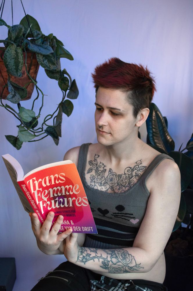 My friend reading page 69 of the book trans femme futures in front of a ficus and a satin pothos