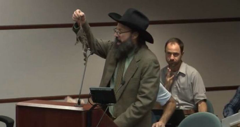man holds up a dead rat at a city council meeting. Person in background looks mildly confused. 