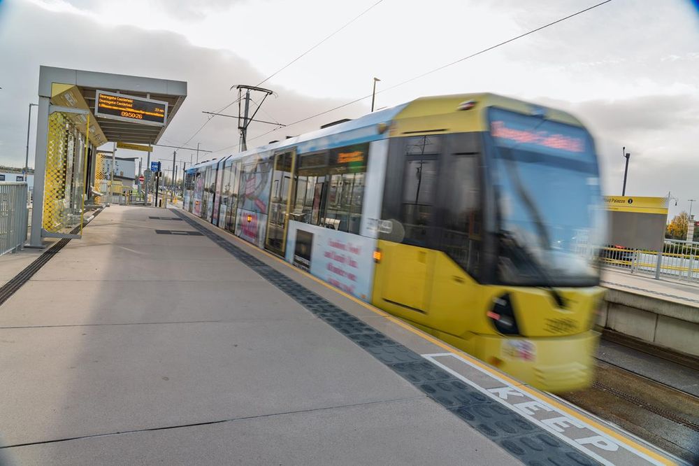 A Metrolink tram in motion at a station with a yellow and black design. The platform features a sign displaying the next tram time and a 'Keep' marking on the ground. The tram is blurred due to its movement, and the station is modern in design.