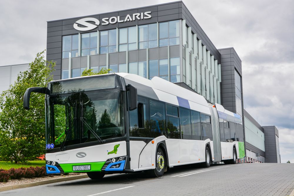 The image features an electric articulated bus parked in front of a modern building with a sleek design. The bus is mainly white with green accents and has a smooth, futuristic shape. The front of the bus displays a logo and a green panel with the word "ELECTRIC" on the number plate area. The building behind the bus is a multi-storey structure with large glass windows and a contemporary façade. The sky is overcast, and there is some greenery, including trees and bushes, visible along the pavement where the bus is located.

Photo by Solaris