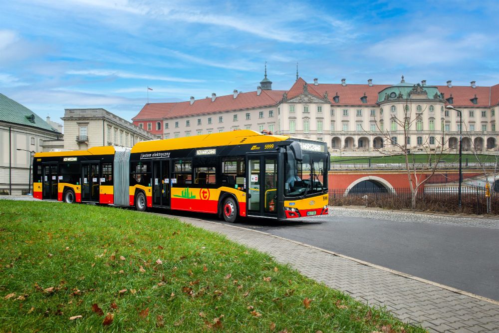 A long, articulated yellow and red Solaris Urbino 18 electric bus drives on a road with a grassy bank to its left, set against a backdrop of a historic building complex with red tiled roofs.