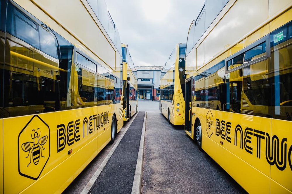 The image shows two bright yellow double-decker buses parked next to each other, with a narrow asphalt lane between them. Both buses have large, bold black text on their sides that reads "BEE NETWORK," accompanied by a logo of a bee within a hexagonal shape. The buses are lined up in a spacious, open garage area with a large industrial building in the background under a cloudy sky. The buses have tinted windows and sleek, modern designs.

Photo by TfGM