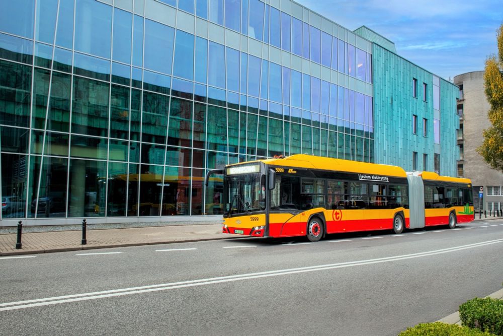 An articulated, yellow and red Solaris Urbino 18 electric bus is parked on a street next to a modern building with large glass panels and turquoise cladding.