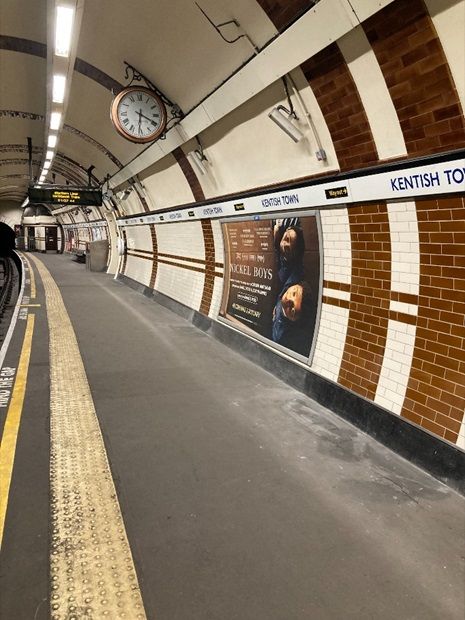 The image shows an Underground station platform with a curved ceiling. The right side of the platform has a wall covered in brown and white tiles, separated into vertical, rectangular sections. A large clock is mounted high on the wall. An advertisement with an artistic design and text is displayed on the tiled wall. Above, a blue sign reading "Kentish Town" is bordered in white. On the left, the train tracks are out of view but run alongside the platform. Overhead lights illuminate the mostly empty platform.

TfL Image - Kentish Town platform