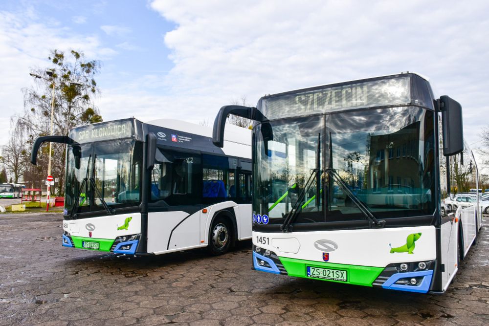 The image features two modern city buses parked side by side in an outdoor setting. The bus on the left has the destination "SPA KLONOWICA" displayed in illuminated text at the front. It is primarily white with black accents around the windows and has a logo and some text on the side. The bus on the right displays "SZCZECIN" as its destination. This bus also has a white body with black window frames and additional green detailing under the windscreen. Both buses have a stylish design with large panoramic windows. The registration plates are visible, with the nearest bus showing the plate "ZS-021SX". The backdrop features a cloudy blue sky and some leafless trees, enhancing the urban outdoor atmosphere.

Photo by Solaris