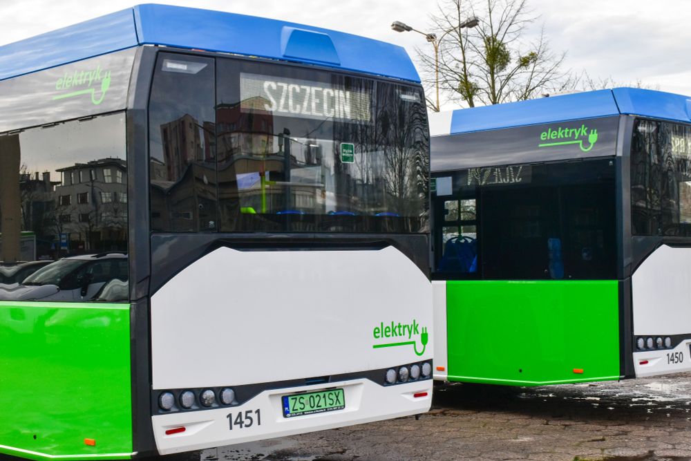 The image shows two electric buses parked side by side. Both buses have a modern design, predominantly painted in white, green, and blue. The rear bus displays "Szczecin" on its destination sign. The rear end of the bus features a digital display panel and several round headlights. The number plate reads "ZS 021SX". The word "elektryk" with an electrical plug icon is visible on the side of both buses, indicating they are electric. The large, dark-tinted windows reflect buildings and cloudy skies. The surroundings include an overcast sky and streetlights.

Photo by Solaris