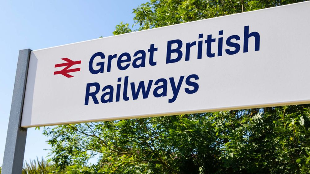 A close-up photograph of a white signpost, typical of a railway station, featuring the Great British Railways logo. The sign displays the red double-arrow symbol next to “Great British Railways” written in navy blue, set against a bright blue sky and green foliage.