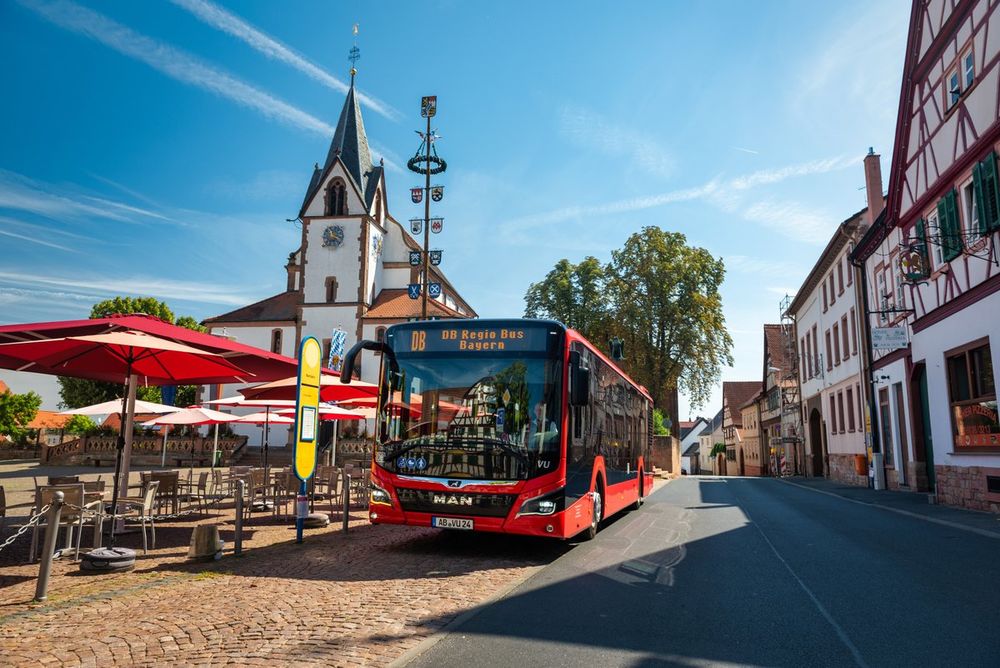 A red MAN Lion's City bus, operated by Deutsche Bahn, is parked on a cobbled square in a small German town. A church with a clock tower and half-timbered buildings are visible in the background under a blue sky.