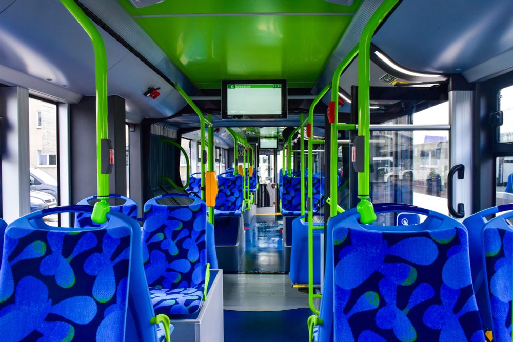 The image shows the interior of a modern city bus. The bus has blue seats with abstract leaf-like patterns in lighter blue and green against a dark background. Bright green poles and handles are positioned for passengers to hold onto while standing. The ceiling is predominantly a vivid green, continuing the vibrant colour scheme. Large, clear windows line both sides, allowing ample natural light to flood the bus, enhancing visibility inside. At the front, an electronic display screen is fixed to the ceiling, likely showing route information.

Photo by Solaris