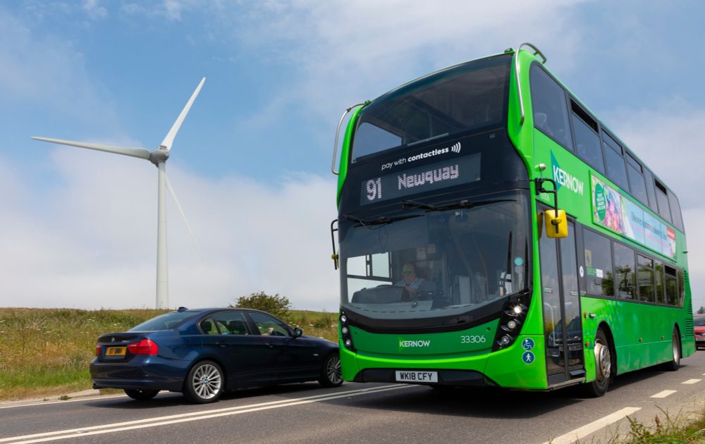 A green double-decker bus on route 91 to Newquay travels along a rural road beside a wind turbine, with a dark blue car passing in the opposite direction.
