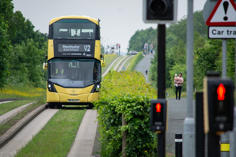 The image shows a yellow double-decker bus travelling along a guided busway, bordered by lush green hedges and grass. The bus displays the destination “Manchester Royal Infirmary” with the service number “V2” on the front. Two red indicators, likely pedestrian crossing signals, are visible in the foreground, with a road sign above showing an exclamation mark and the words “Car trap.” In the distance, a person is walking carrying a child along a path parallel to the busway. The scene is surrounded by dense trees and vegetation beneath an overcast sky.

Photo by TfGM