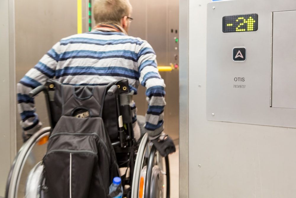 A person in a wheelchair, seen from the back, is facing a lift car and reaching for the control panel to select a floor. They are wearing a blue and white striped long-sleeve shirt. A black rucksack is attached to the back of the wheelchair, with a blue water bottle in a side pocket. The lift display shows "−2" in yellow-green lights. The lift panel has an OTIS logo and an arrow pointing up on the main button.

Photo by TfL