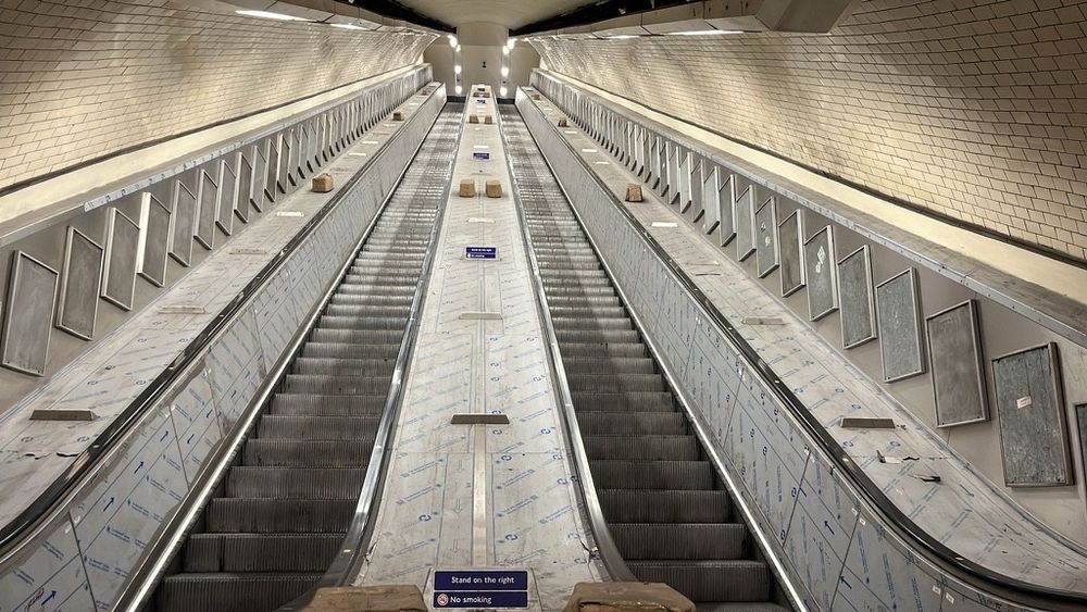 The image shows a pair of parallel escalators in a tube station, extending upwards into the distance. The escalators are out of use, covered with white protective sheeting bearing blue printed patterns and arrows. The surrounding walls are lined with cream-coloured, glossy tiles in a grid pattern, which curve overhead. The escalator side panels contain metallic frames at an angle. Bright overhead lights illuminate the scene. At the base of the escalators, there are signs and small blocks on the steps.

TfL Image - Kentish Town escalators