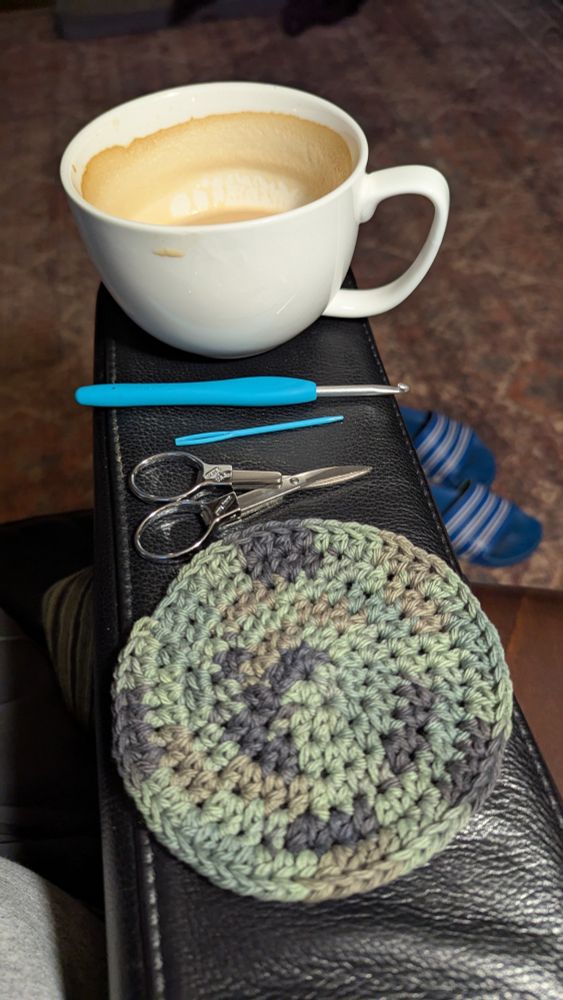This is a picture of my morning crochet work. Top to bottom: white coffee mug, crochet hook, needle, small scissors, crocheted coaster in various shades of green