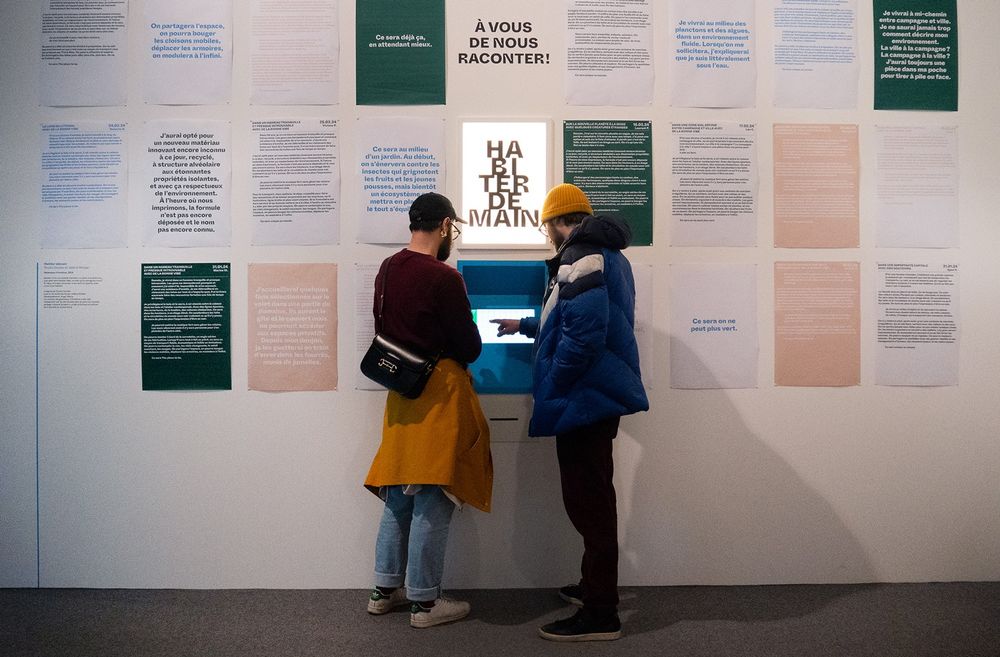 people standing in front of the "Habiter Demain" story generator, in the Tracé Bleu exhibition in CENTQUATRE-PARIS. there is a touch screen inside the wall, and that wall is covered in posters of generated stories.