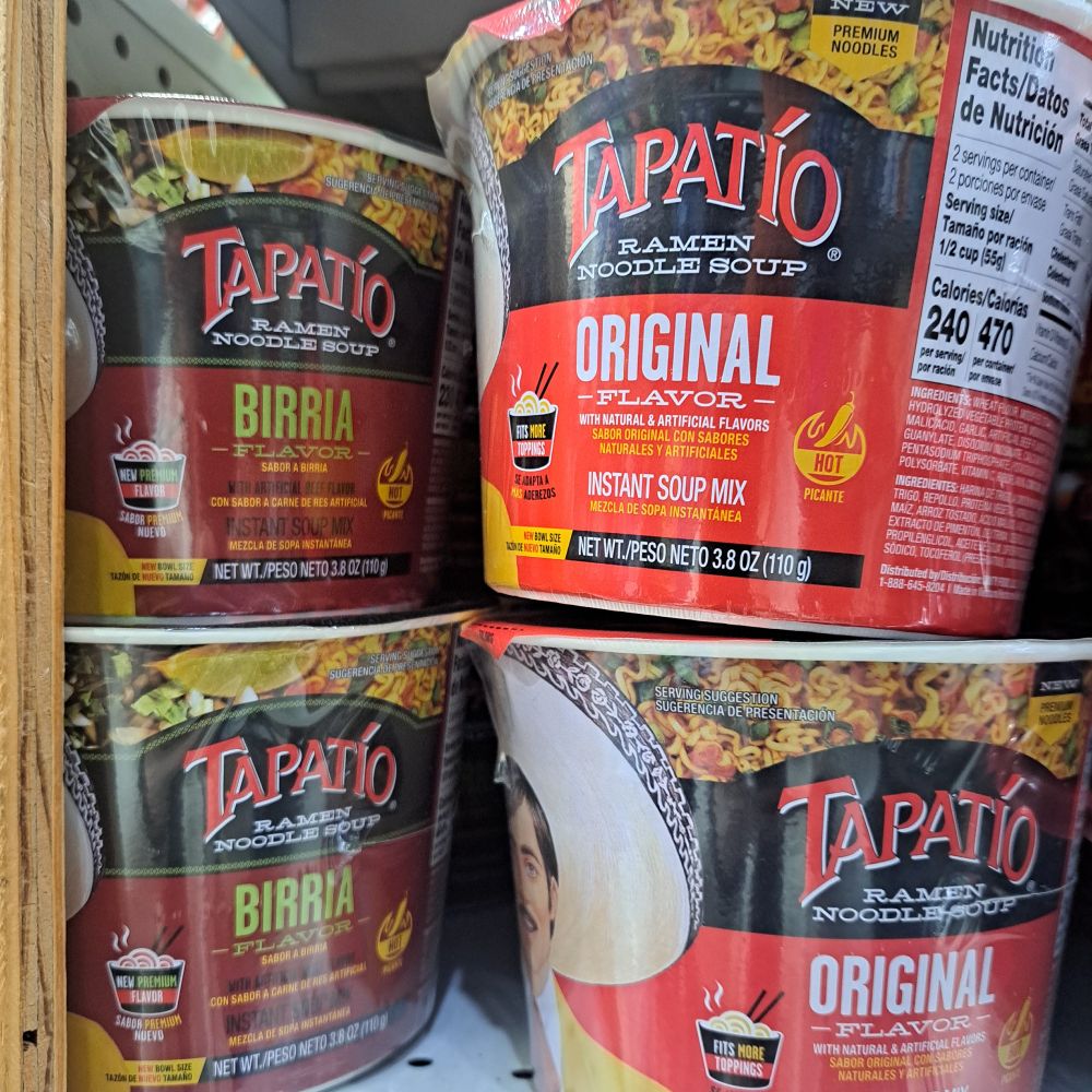 Photo of a shelf from my corner deli which holds two kinds of Tapatio ramen bowls, regular and birria