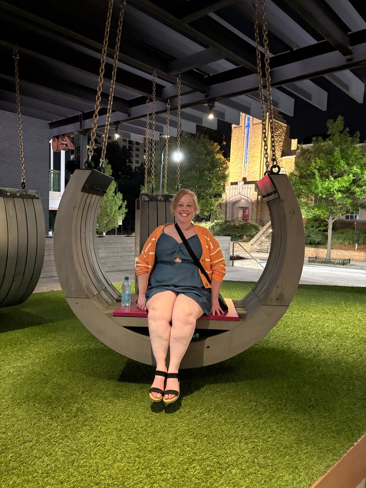A cis white female dressed in a denim romper with an orange and striped cardigan, sits in a large half circle swing smiling at the camera.
