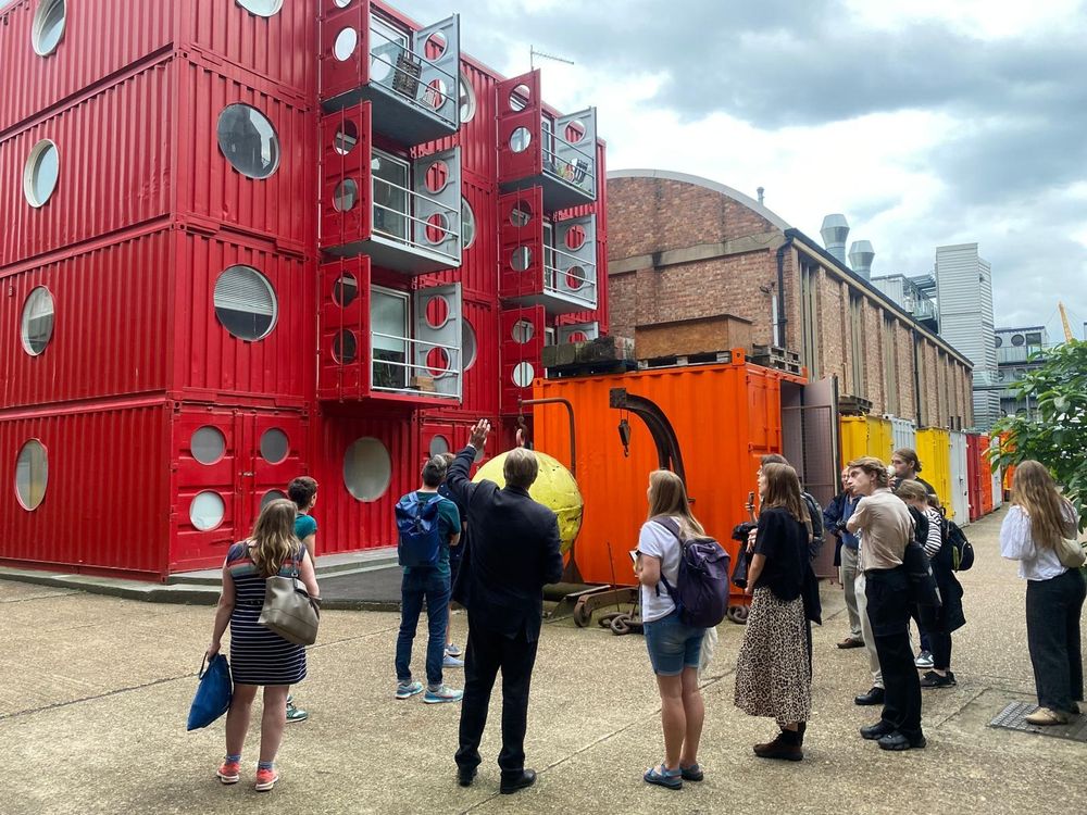A group of people on a walking tour. They stand in front of one modern, container style building and an older brick built building. 