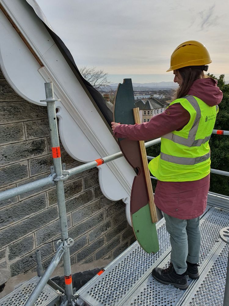 Photograph of a woman standing on scaffolding in a hard hat and high-vis jacket next to the gable end of a building.