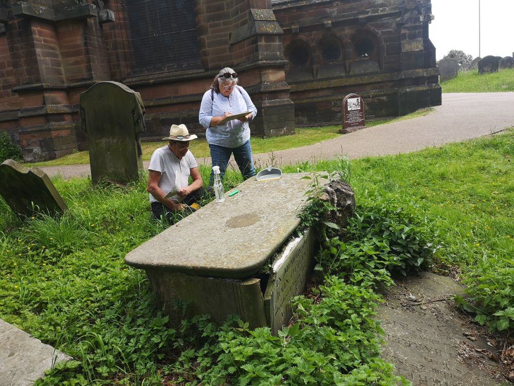 Two people next to a gravestone.  They are clearly studying it and the woman is writing something on a clipboard. 