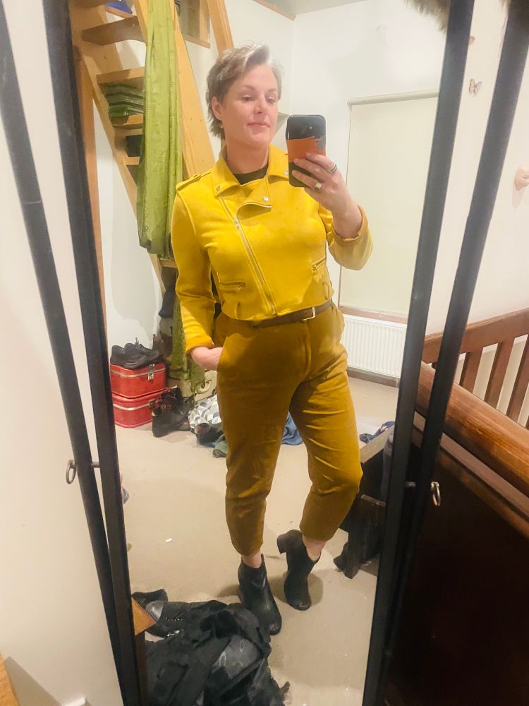 Me a woman with grey short hair is wearing tan pants that stop at the ankle
Black ankle boots that have a small heel and a funky yellow suede bolero jacket and its a fine look