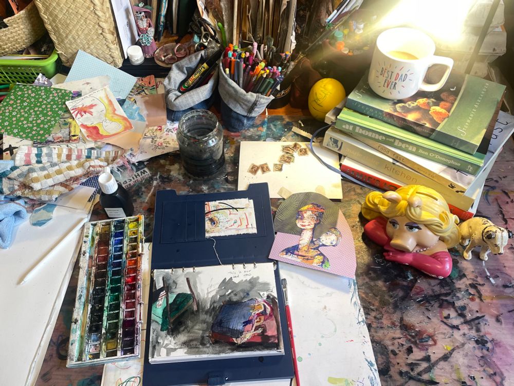 A cluttered desk witb piles of paper and drawings and miss piggy and also a his masters dog moneyboxes
A pan of square watercolour paints 
And a sketch of beds in the centre 