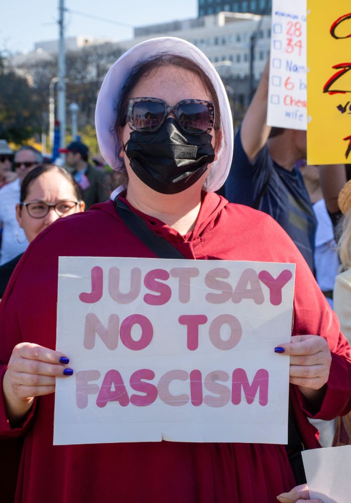 This image shows a woman dressed like she's from A Handmaid's Tale holding a sign that says "Just say no to Fascism."