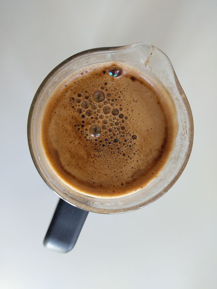 Blick von oben auf eine French press ohne Deckel, zu sehen ist der duftende Kaffeeschaum in unterschiedlichen Braunnuancen. In der Mitte und am Rand sind einzelne schillernde Bläschen zu sehen.
