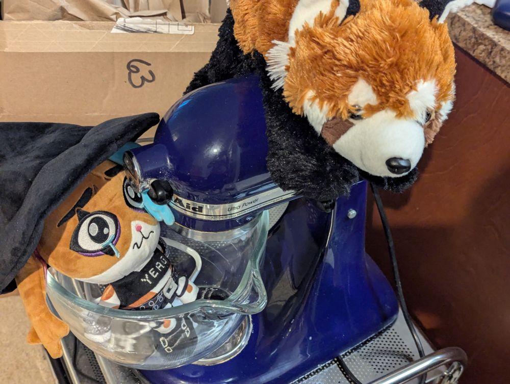 Bunny plushie in mixer bowl and red panda plushie with paw on mixer's on/off switch 