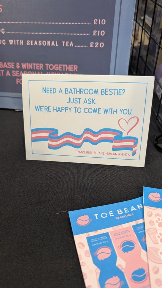 Table sign that reads: Need a bathroom bestie? Just ask. We are happy to come with you. Trans rights are human rights. There is a ribbon coloured with the trans flag and a heart.